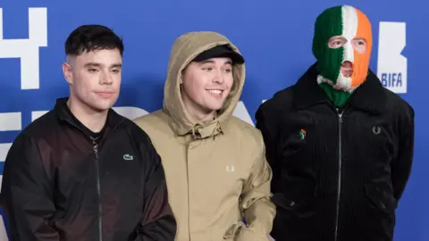 Getty Images Kneecap members Moglai Bap in a black bomber jacket, Mo Chara in a beige hooded jacket and black baseball hat and DJ Provai in a black bomber jacket and green white and orange Irish tricolor balaclava attend the British Independent Film Awards 