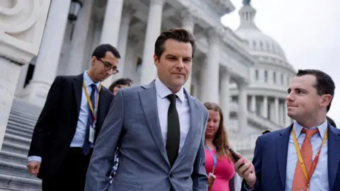 Reuters Matt Gaetz in grey suit with dark tie, before white marble buildings, with reporters around him