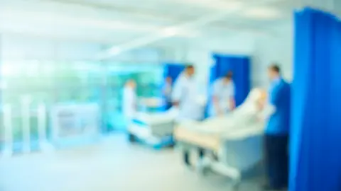 Getty Images A blurred image of what appears to be a hospital ward with medical staff in blue uniforms moving between beds. The image is intentionally out of focus, giving a sense of constant movement and activity.