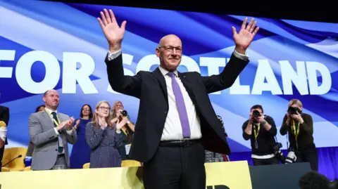 Getty Images John Swinney at an SNP conference, arms aloft, taking applause in front of a big sign reading 