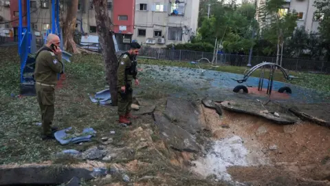 Reuters Two Israeli military personnel wearing green combat gear examine a crater left by a Houthi missile attack in Tel Aviv. In the background are swings and other playground apparatus.