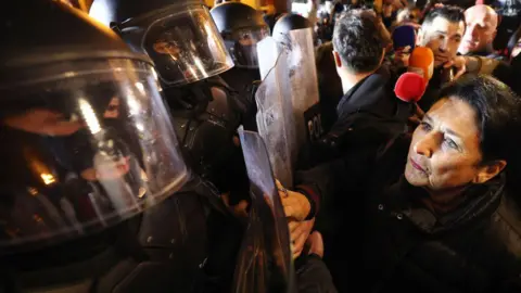 Getty Images Salome Zourabichvili facing a row of masked riot police. The riot police are wearing balaclavas under riot helmets and holding riot shields. A crowd surround the president. 