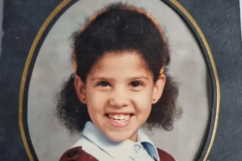 Anastasia Cameron Anastasia is in her primary school uniform, posing for the professional pictures taken in school. Her curly, black Afro hair is cut to chin length and a hairband is on top of her hair.