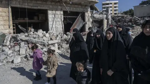 Getty Images Two little girls in coats hold hands as they walk along a street filled with rubble with a group of women who are wearing hijabs 