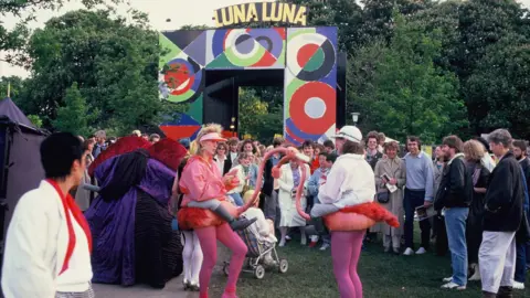 © Sabina Sarnitz. Courtesy Luna Luna, LLC Two performers dressed as flamingos touch beaks at the center of a crowd.