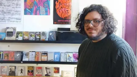 Alex Tadross Alex Tadross stands in frong of shelves of cassette tapes