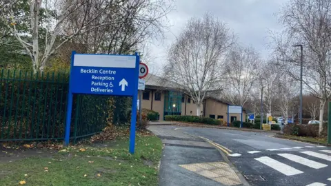 BBC/Sabbiyah Pervez The outside of the Becklin Centre, which is a low, brown-brick building surrounded by trees. A sign pointing visitors towards the car park and reception is bright blue and sits to the left of the road and pavement leading up to the building.