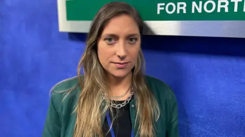 BBC Fernanda Hermosilla standing in front of an Equality Commission for Northern Ireland sign.  She has long, straight, dark-fair hair and is wearing a green jacket over black top and two silver necklaces. 