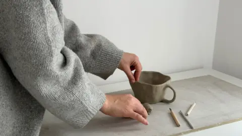 Mollie Warrington An image of a woman's hands working on an unfired piece of pottery in the shape of a cup, alongside some tools on a bench