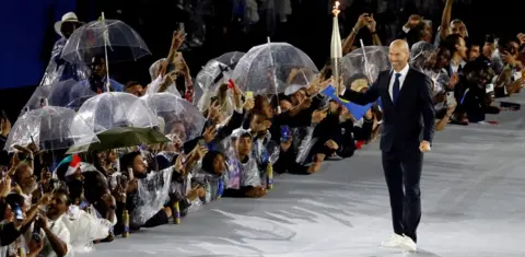 Reuters French former football player Zinedine Zidane carries the Olympic torch at the Trocadero during the opening ceremony of the Paris 2024 Olympic Games