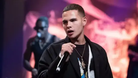 BBC Slowthai performing holding a microphone with a light orange and purple background