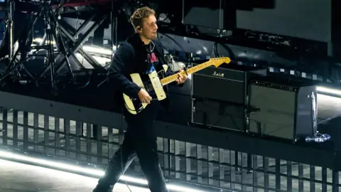 Getty Images Sam Fender on stage with a black and white striped electric guitar. He's walking with a smile on his face.