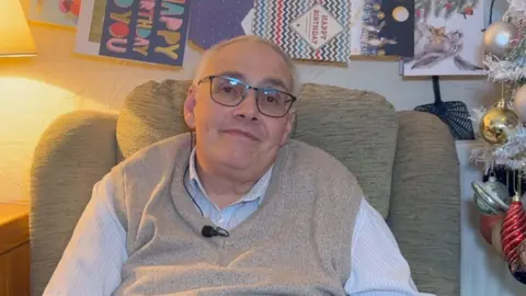 Gary Jameson sitting in a grey armchair with birthday cards behind him and Christmas baubles on the right of him. 