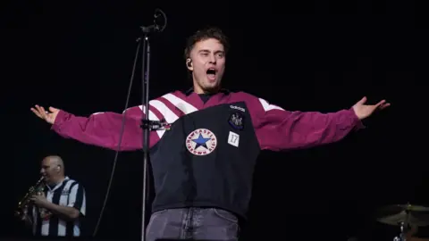 PA Media Sam Fender on stage with his arms outstretched, shouting. He's wearing a black and maroon vintage Newcastle United top.