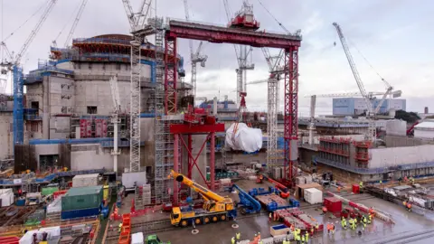 Hinkley Point C A big cylinder shrouded in white wrapping is lifted up on a big red crane. It stands in front of a large concrete building, with a steel brown domed roof. Dozens of workers in high vis are on the ground, and there are several other cranes all around.