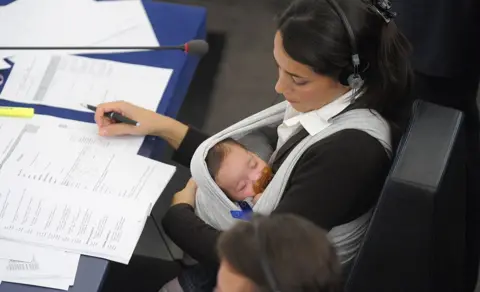 Getty Images In a photo from 2010, European deputy Licia Ronzulli sits in parliament and signs papers while she holds her baby who is asleep in a sling, sucking a pacifier