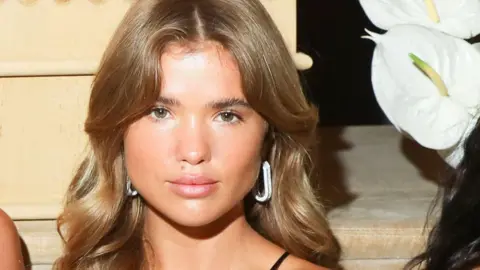 Shuttershock Matilda Djerf with long light brown hair and silver earrings on, with a white flower seen next to her