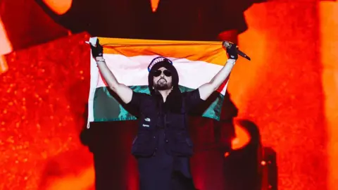 X/diljitdosanjh A picture of artist Diljit Dosanjh wearing a black Indian traditional shirt with a black jacket, sunglasses and a black turban holding the Indian flag and a mic during his concert in Delhi.