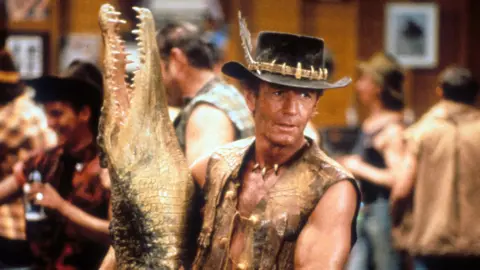 Getty Images Paul Hogan with fake dead crocodile while filming crocodile Dundee. He is wearing a hat and waistcoat.