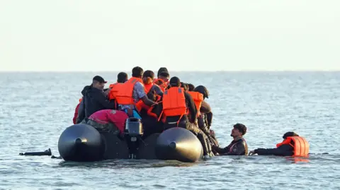 PA Media A group of people thought to be migrants wade through the sea to clamber aboard a small boat off the beach in Gravelines, France