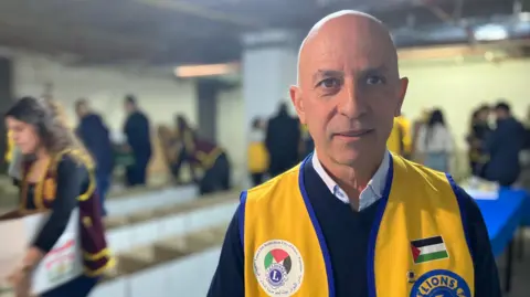 Wael Shaer stands looking directly at the camera, wearing a yellow charity vest, inside a busy room with people in the background packing boxes