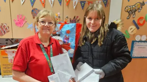 TEAM MIKAYLA Mikayla Beames poses with an unnamed hospital ward staff member. They are both holding what appear to be Christmas presents and smiling in front of the ward's cupboards which are decorated with bunting and children's drawings.