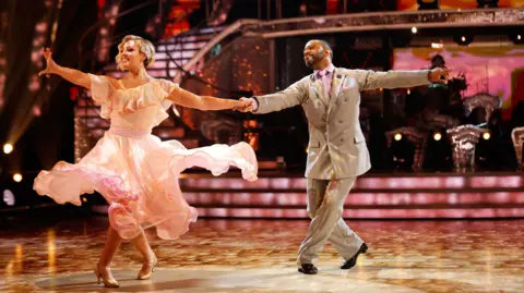 Amy Dowden and JB Gill, during an appearance on Strictly Come Dancing in September. Dowden is wearing a light pink frilly dress and dancing shoes and Gill has on a grey suit and pink tie. They are performing and holding hands as they dance together with their arms outstretched.