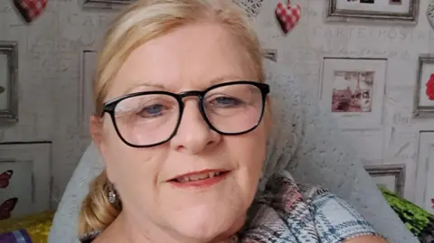 Wendy Swain Wendy Swain at home. She has glasses on and is smiling.  She has blonde hair, pulled behind her head in a ponytail and has blue eyes. Her mouth is slightly open. Behind her on a wall decorated with wallpaper with grey writing, are a series of framed pictures.