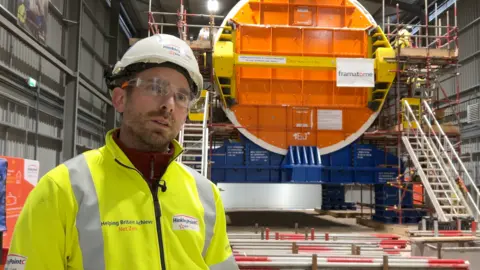 Matt Abbott stands in front of the huge reactor in its storage warehouse. He is wearing yellow hi-vis jacket and a white hard hat. The end of the cylindrical reactor unit is behind him, with a bright orange disc at the front.