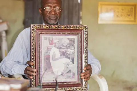 Ifiokabasi Ettang / BBC Bala Muhammad, wearing wire-rimmed glasses and a long-sleeved pin-stripped blue shirt, sits in a white-painted wooden chair holding up a wide-wooden framed black-and-white photograph of his father, who is pictured looking at the camera as he sits at his workshop bench. He is wearing a sleeveless boubou. A desk fan can be seen in the background and a pendulum clock hangs on the wall