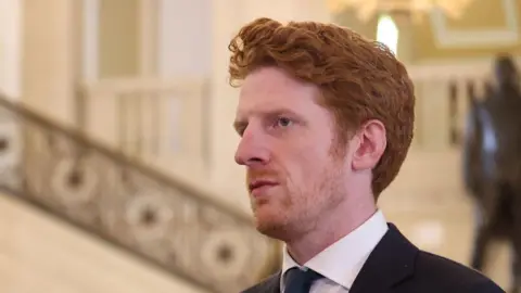 PA Media Matthew O'Toole looking sideways with the staircase of the great hall at Stormont in the background. He has red hair and wearing a black blazer and white shirt and dark green tie.