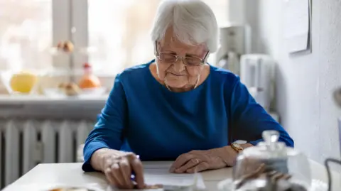 Getty Images An elderly woman sitting at a table looks at a form