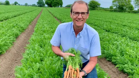 Rodger Hobson smiling man in a blue shirt and glasses, kneeling in a carrot field and holding up some freshly dug-up carrots.