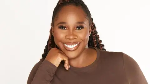 Amanda Akokhia Denise is wearing a brown top and is pictured against a plain white background. She has long, black braids with curls and has her chin resting on her hand and is smiling.