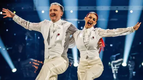 BBC Chris McCausland and Dianne Buswell dancing on Strictly in matching white  suits, smiling and arm in arm, each with their other arm outstretched and with a leg raised.