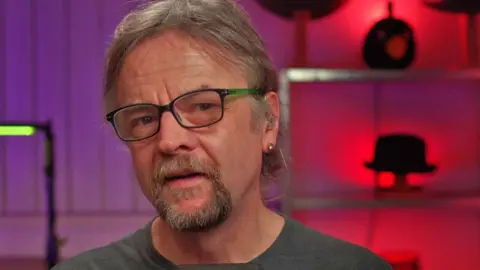 Paul Duckett Paul Duckett wearing glasses and a grey T-shirt. The wall behind him is lit purple and red