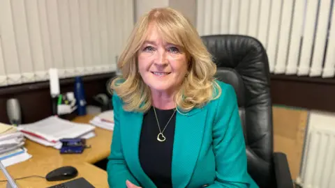 Geraldine McGahey sitting at a computer desk.  She has shoulder-length, curled blonde hair and is wearing a bright green blazer, black top and a gold heart-shaped necklace. 