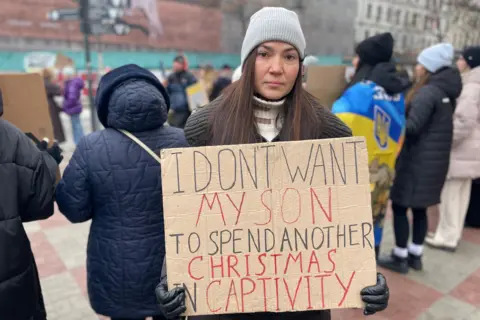 A woman is seen holding a cardboard sign which says 