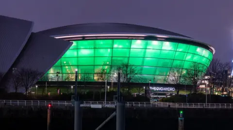 Getty Images A large events arena with its  windows lit up in green in front of a river