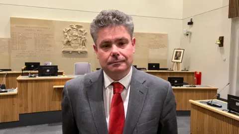 Simon Dedman/BBC John Kent wearing a white shirt, red tie and grey suit jacket. He has grey hair. He is standing in the council chamber.