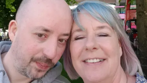 Roisin Wilshaw Brian is pictured in a selfie with his mother Isobel, Brian is bald and goatee and is wearing a grey hoodie. Isobel has a blue fringe and pink highlights on her short light hair. They are standing outside near buses.  