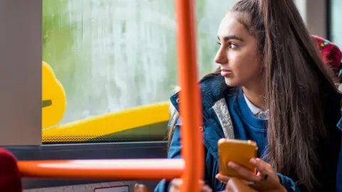 Getty Images Teenage girl wearing a ponytail and a navy blue parka coat sits on the bus while holding her smartphone in her left hand and gazing out the window