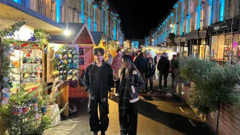 BBC Man and women walking through a Christmas market at night, with stalls selling Christmas gifts and Christmas trees and lights running along the side.