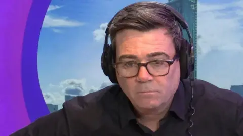 BBC Andy Burnham wearing a black shirt and headphones, wearing glasses, in a radio studio. He is looking pensively towards the camera. 