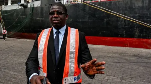 AFP Abidjan port boss Hien Yacouba Sie speaks to reporters. He is wearing a hi-viz tabard. Behind him in the outer harbour is the Zimrida vessel.