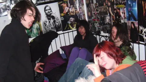 AmberWillis/MuseumOfYouthCulture Four people on a black metal frame bed surrounded by posters of emo bands on the wall. A boy on the left has long brown hair and a black jacket, in the middle is a girl with black hair and a fringe. She has a black hoodie and purple trousers. To the right of her is a boy with long hair wearing a camouflage top and jeans. In the front right is a person with red hair motioning a peace sign with her fingers. 