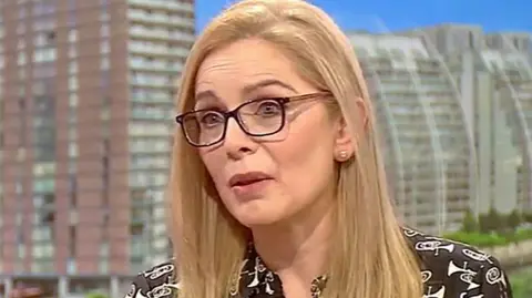 Dr Kalena Marti with long blond hair wearing a dark brown top with a pattern of white trumpets with the views of Salford Quays in the background