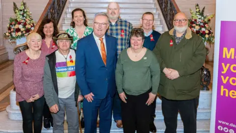 Eight members of Dementia NI stand at the bottom of a large staircase at Stormont. They are wearing red ribbons and smiling at the camera alongside Mike Nesbitt. He is wearing a blue suit with a white shirt and orange tie and is also smiling at the camera.