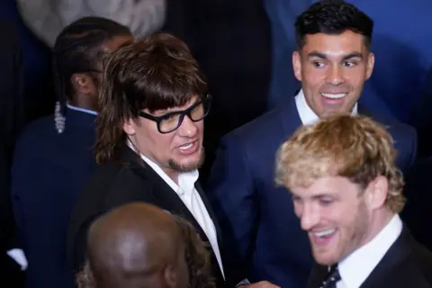 Getty Images A man with a mullet and glasses stands next to a man with dark hair and a man with blond shaggy hair. They all wear suits.