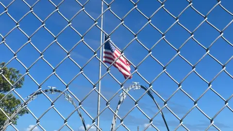 BBC An American flag flies against a blue sky behind a chainlink fence and barbed wire at Guantanamo Bay